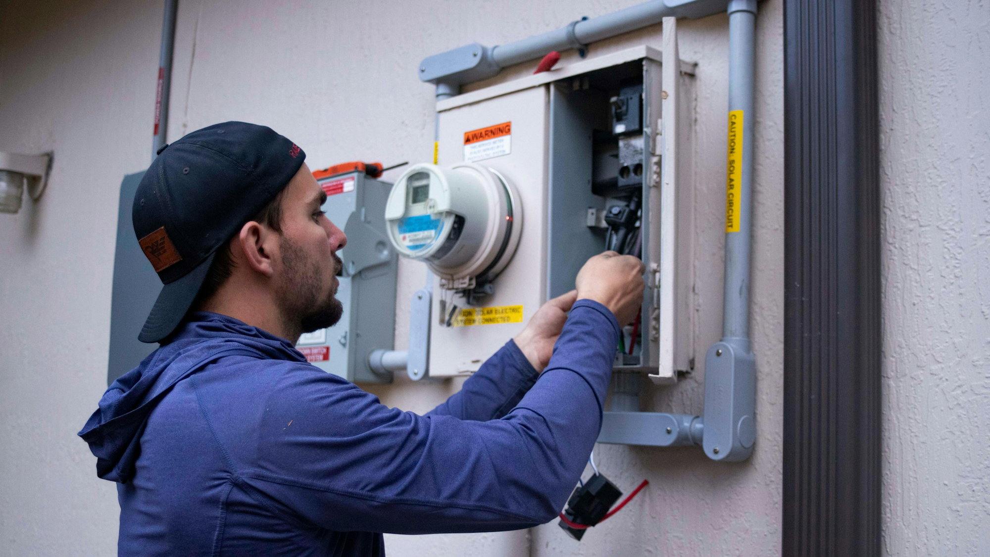 A person in a blue hoodie and baseball cap working on an electrical meter